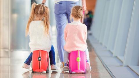 Two young girls sit on kid-sized luggage suitcases known as a Trunki.
