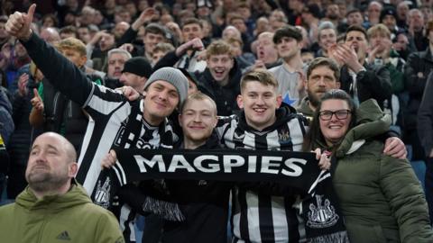 Newcastle fans hold a scarf saying "Magpies" at Villa Park. They are wearing black and white striped shirts.