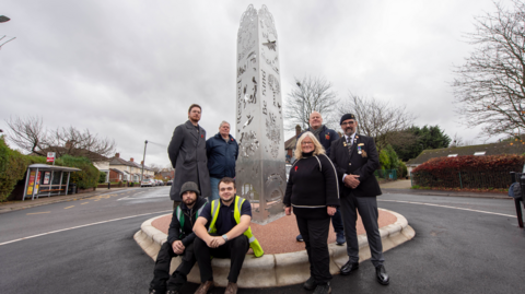 A group of seven people are in front of a metal sculpture, which looks to be in the middle of a roundabout. On the left of the sculpture two of the men are sitting, while two others stand behind them. To the right two men and one woman are all standing