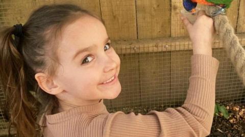 Aria Thorpe. She is a young child with long, dark hair tied up in a ponytail. She is pictured feeding a colourful bird and is looking over her shoulder to smile at the camera.