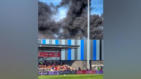 The image shows thick black smoke billowing into the sky behind a football stadium during a match. Spectators are visible seated in the stand below, with advertising boards along the pitch and a tall floodlight pole in the foreground. The stadium exterior features blue and grey metal cladding.