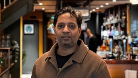 Raja Munuswamy, who wears a brown jacket and has dark brown hair and a short beard, poses for a picture in a restaurant. A bar is visible behind him.