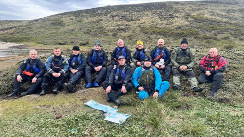 Image shows 11 men sitting on a green hillside, dressed in waterproof clothing and life jackets. They are all looking at the camera. 