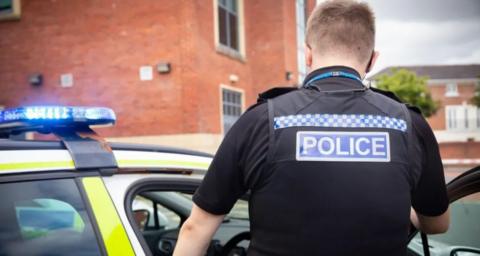 A police officer wearing a black uniform is facing away from the camera and getting into his police car. The vehicle is parked on a residential road next to a tall red brick building.