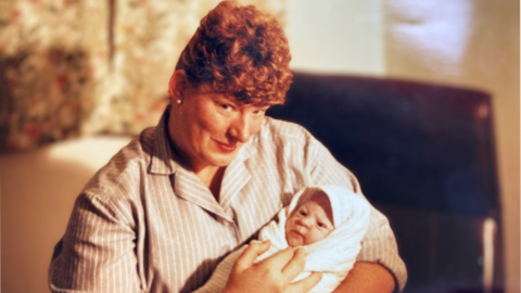 Diane Hele in 1989 smiling at the camera while cradling a newborn baby wrapped in a white blanket. 