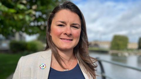 Charlotte Horobin with long brown hair, dark blue top and checked jacket, standing outside in front of the River Nene.