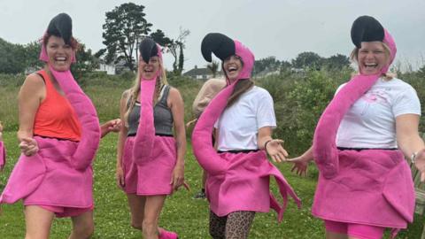 Four women dressed as flamingos posing for the camera. They are all holding out their hands and smiling
