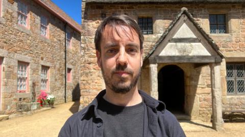 Ben Spink stands centered in a sunlit stone courtyard, facing the camera, wearing a dark shirt and jacket. Behind him are rustic buildings made of warm-toned stone, including one with pink-trimmed windows on the left and another with a thatched roof and a small wooden porch on the right. Bright blue sky and a pot of red and white flowers add colour to the scene.