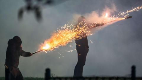 Two masked youths weaing black trousers and hoodies set off fireworks from their hands, sparks fly and the rockets launch in a smokey night time scene