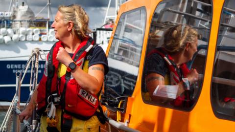 Sarah Berrey is standing next to an orange RNLI lifeboat, looking into the distance while wearing her lifeboat safety uniform and red life vest 