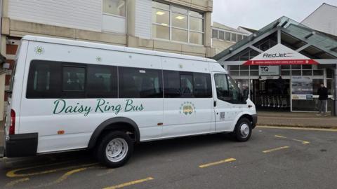 A white minibus with 'Daisy Ring Bus' in green text written along side, parked on a bus stop outside the RedJet terminal doors