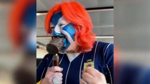 Michelle McManus wearing Saltire face paint and a Scotland football top, speaking into an old microphone