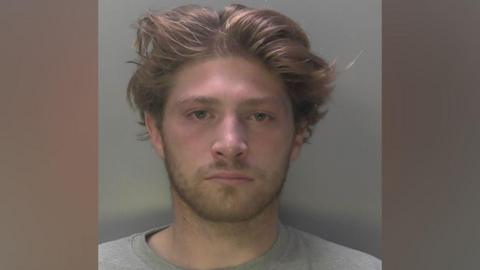 A police mugshot of a man with long hair in a grey top.