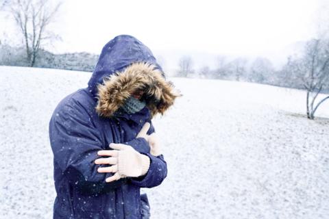 A person stands on hard, snowy ground shivering, wrapped in a winter coat, scarf and gloves.