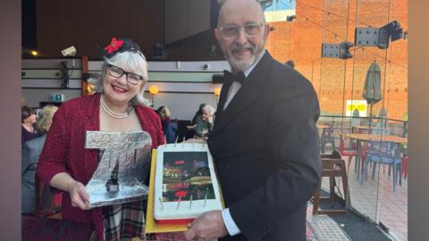 A husband and wife, probably in their 70s in a pub. The man is holding a birthday cake with a picture of a pub at night on it, and the woman is holding a big number "70" made out of silver foil. She is wearing a red and black blazer, floral headpiece, and black glasses, and he is wearing a black and white suit and bowtie. Other people can be seen sat behind them inside, and there is an outdoor seating area lit up by sun on the other side of large panes of glass.