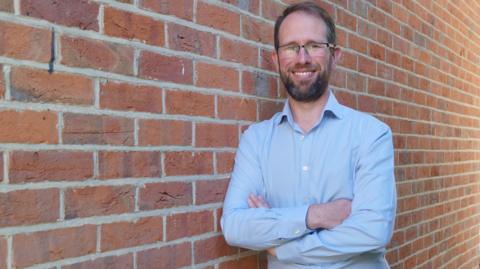 Matthew Barber, Police and Crime Commissioner for Thames Valley, wearing a blue shirt and standing against a brick wall with his arms crossed.