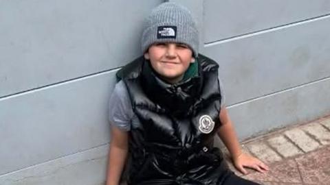 Coby Keogh sits on red brick paving leaning against a grey fence. He is smiling and wearing a shiny black gillet and grey beanie