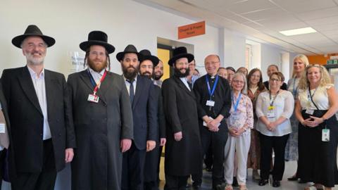 A group of Jewish community leaders and NHS staff standing by the door of the new prayer space while smiling.