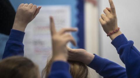 School children in a classroom. They have their hands up and only their hands, arm and top of their heads are visible. They are wearing blue jumpers and one of the children has a bracelet on.
