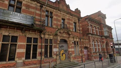 The building has red bricks and ornate stone surrounds to the windows. The front entrance and window to the right of it has been boarded up and has graffiti on it. The sign saying East Hull Pools is still on the front to the left of the entrance.