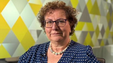 Dame Hilary Cass is sat looking at the camera in a meeting room. She is in front of a glass wall with a geometric pattern.