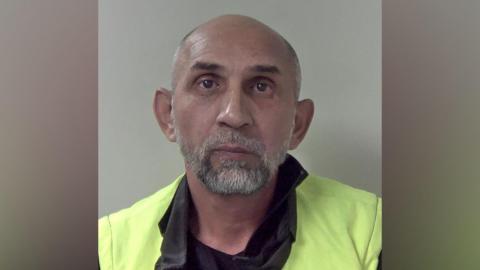 A bald man with a beard wearing a neon yellow jacket in a police mugshot.