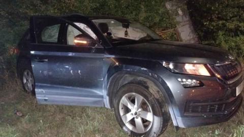 A car resting on grass at the side of the road with the lights still on and clear damage to its doors
