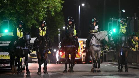 A line of police on horseback face towards the camera at night time in Bristol. In the background lights of police vehicles can be seen