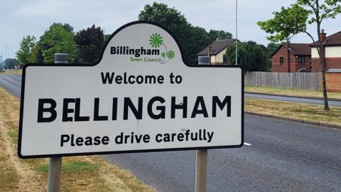A black and white road sign that now reads: "Welcome to Bellingham. Please drive carefully." There is a grey tarmac road behind with a grassy verge. There are residential houses and lush green trees beyond it.