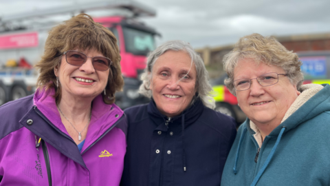 Three women wearing jackets posing together for a photo. The large vehicles are seen in the background.
