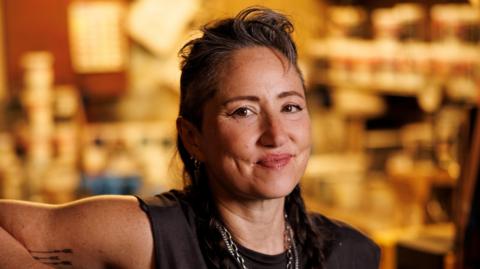KT Tunstall in black T-shirt and necklaces, with her hair swept back off her face. She is smiling. The gold-coloured backdrop is out of focus.