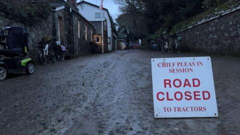 A sign ona. muddy floor which says Chief Pleas in session, road closed to tractors. The writing is red on a white sign. Along the wall are a few bikes and a mobility scooter. It's dusk, so the light is dull. 