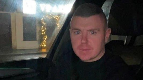 A man with short fair hair and wearing a black top is sitting in the front seat of a car