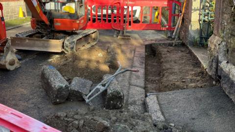 Excavated pavement in front of church gate with kerb stones pulled out and bare ground. Some tools and digger and bollards to prevent people walking there