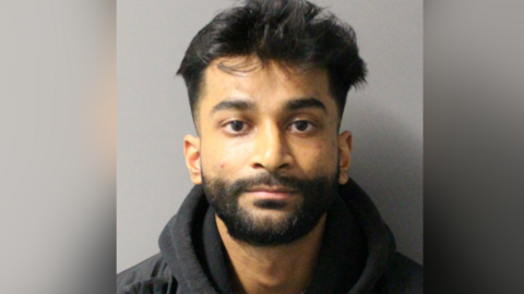 A man looks at the camera for a police custody photo. He has a dark brown beard and brown hair and is wearing a black hoodie