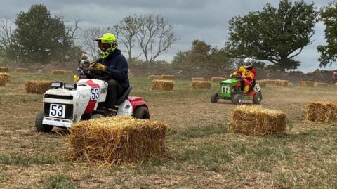Two men on lawn mowers with racing colours and numbers racing down a a dirt track. 