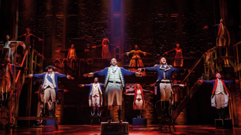 Hamilton actors on stage wearing blue long military jackets, white trousers and black hats. They are singing with their arms stretched out. There is a red shine on some of the actors and a few women in grand ball dresses are stood in the background.