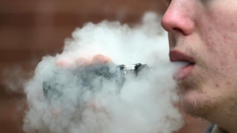 A man holding a disposable vape exhales a cloud of vapour