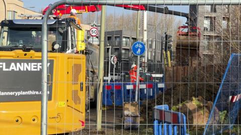 Metal fencing with machinery behind and plastic fencing with worker in orange high vis and white helmet.