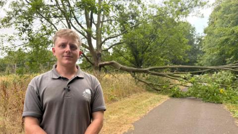 A man standing in the foreground, wearing a grey polo top and looking at the camera. In the background, a large branch has fallen off a tree and is blocking the road.