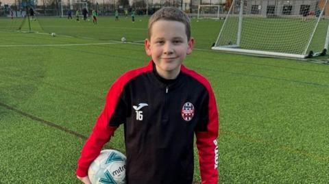 Harry smiling wearing a red and black football strip holding a football on a pitch.