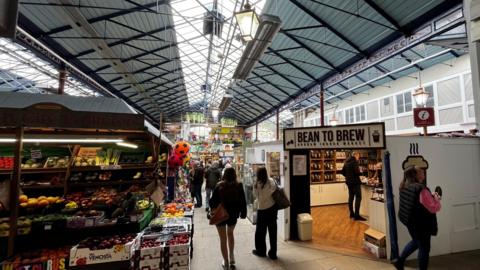 People stroll along the market which is full of stalls, including fruit and veg and a coffee shop. 