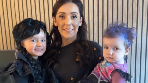 A woman with long brown hair carrying two children, both in halloween costumes