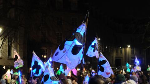 A dark image of Maidstone town centre with brightly lit lanterns being carried on sticks. The lanterns are white with black and red designs, they are being carried by people. 