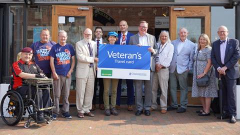 A group of people outside the Soldiers of Gloucestershire Museum. The people in the centre of the image are holding a large blue sign which reads 'Veteran's Travel Card'. (l to r) Veterans Mike Lancaster, Carl Askew, Chris Auker-Howlett, Cllr John Bloxsom, Lee Cassidy, Cllr Cate Cody, Steve Olczak, Cllr Mark Hawthorne, Cllr Colin Hay, Cllr Andrew Gravells, Gill Knight and Cllr Philip Robinson. 