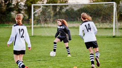 Three teenage girls playing football. The player in the middle is wearing a black kit, including a waterproof jacket and is dribbling the football. Two players on the opposing team, wearing white kits, are heading towards her on either side.