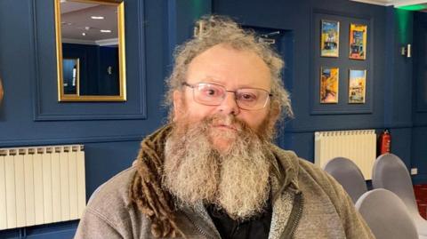 Lance Vaudin He has long grey hair and a grey beard. He is wearing glasses and a grey hooded jumper. 