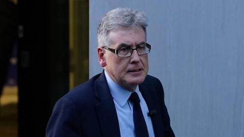 A man with short, grey and white hair, wearing metal-rimmed glasses and wearing a dark blue jacket over a light blue shirt with a dark blue tie, walks from left to right.