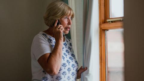 an older woman wearing a t-shirt and holding a phone to her ear clutches a curtain nervously as she looks out of a window