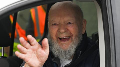 Michael Eavis smiles and waves at the camera from the window of his van